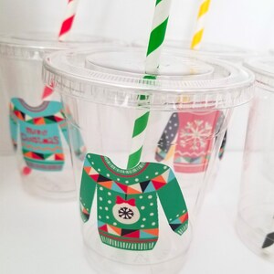 May include: Clear plastic cups with festive Christmas sweater designs. The cups have green, red, blue, white, and black patterns. Each cup has a straw.