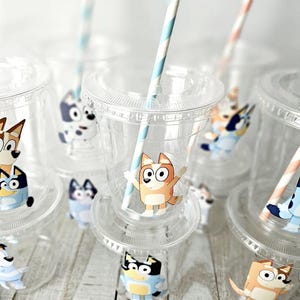 May include: Clear plastic cups with lids and straws, decorated with cartoon dog characters. The cups have a clear design, and the straws are striped in orange and white or blue and white. The cups are arranged on a light-colored wooden surface.
