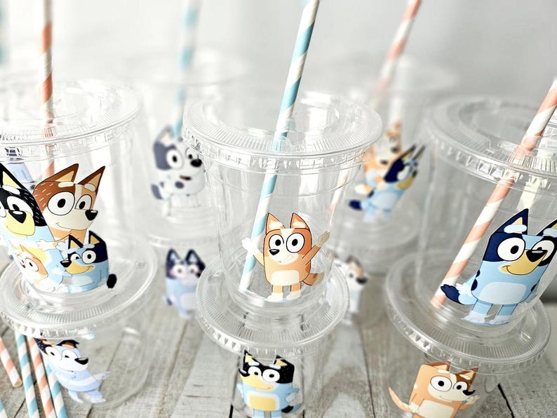 May include: Clear plastic cups with lids and straws, decorated with cartoon dog characters. The cups have a clear design, and the straws are striped in orange and white or blue and white. The cups are arranged on a light-colored wooden surface.