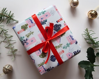 May include: A rectangular gift box wrapped in white paper featuring a repeating pattern of pink houses, red telephone booths, and green pine trees. The package is tied with a vibrant red satin ribbon tied in a bow at the center of the box.