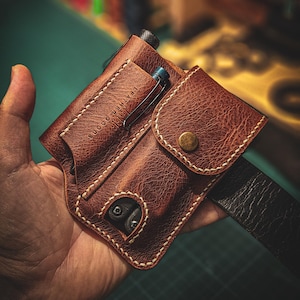 Puede incluir: Una funda de cinturón de cuero marrón con costuras blancas y cierre de botón a presión. La funda tiene un bolsillo para un bolígrafo y una pequeña herramienta. Se ven las palabras "LEATHER GOODS".