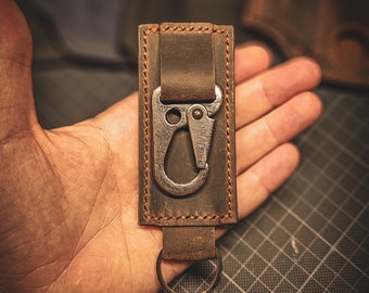 May include: A brown leather key chain with a metal hook and a ring. The key chain is stitched with brown thread.