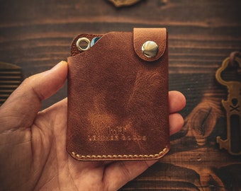 May include: A brown leather wallet with a snap closure and the text "10PM Leather Goods" embossed on the front. The wallet is being held in a hand, and a silver coin with a Spartan helmet design is visible in the background.