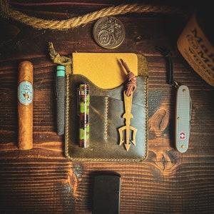 May include: An assortment of everyday carry items on a wooden surface. Includes a cigar, leather wallet, pen, flashlight, pocket knife, lighter, and a brass trident-shaped keychain. The items are arranged in a flat lay composition.