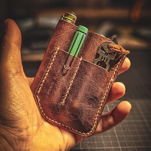 May include: A brown leather pocket organizer with visible light-colored stitching. The organizer features two vertical slots containing a light green pen and a metallic skull-shaped tool with a leather cord attached to the top edge.