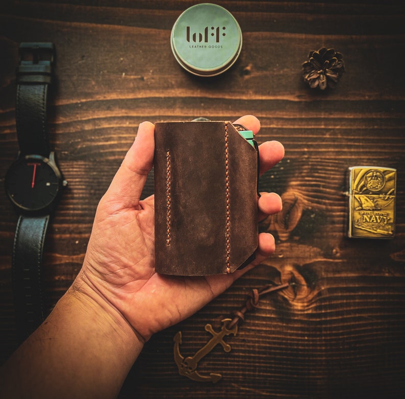 May include: A brown leather wallet with a stitched design. The wallet is being held in a person's hand. The wallet has a small label that says "lofi: leather goods".