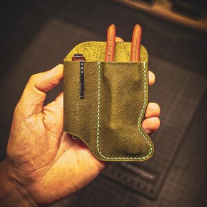 May include: Olive green leather pen holder with a light green stitched border. The holder contains two orange pencils and a silver clip. The text "LEATHER GOODS" is visible on the clip.