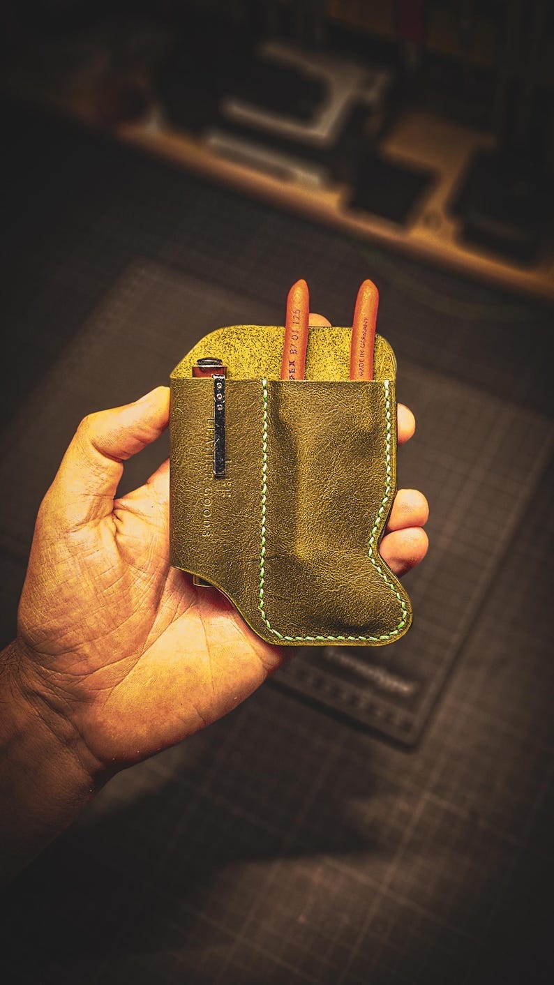 May include: Olive green leather pen holder with a light green stitched border. The holder contains two orange pencils and a silver clip. The text "LEATHER GOODS" is visible on the clip.