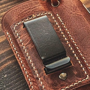 May include: Close-up of a brown leather wallet with a black metal money clip. The wallet features white stitching and a small metal rivet. A leather loop is attached to the top.
