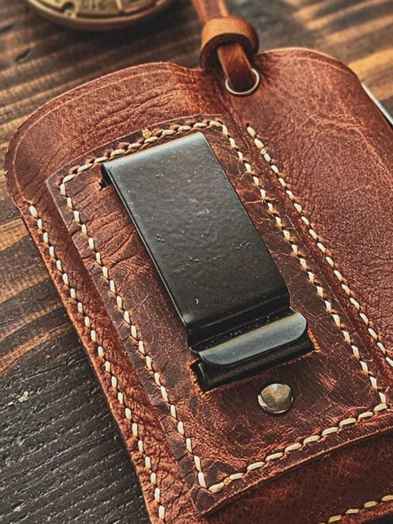 May include: Close-up of a brown leather wallet with a black metal money clip. The wallet features white stitching and a small metal rivet. A leather loop is attached to the top.