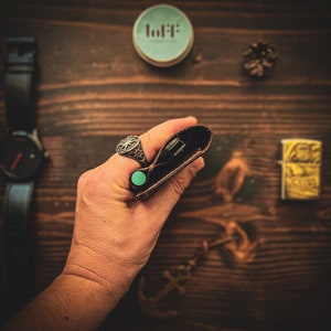 May include: A brown leather wallet with a turquoise button closure. The wallet is being held in a hand wearing a silver ring with a cross design.