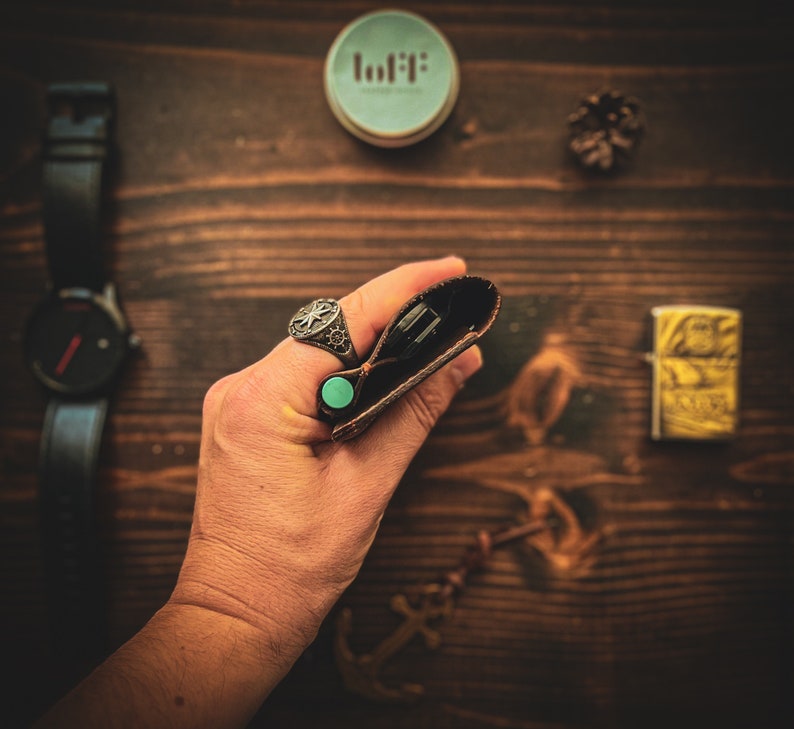 May include: A brown leather wallet with a turquoise button closure. The wallet is being held in a hand wearing a silver ring with a cross design.