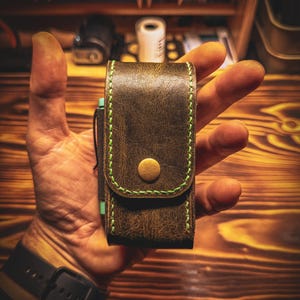 Könnte beinhalten: Eine dunkelbraune Tasche aus Distressed-Leder mit leuchtend hellgrünen Nähten an den Kanten und einem einzelnen messingfarbenen Druckknopfverschluss an der Frontklappe. Das rechteckige Etui ist vertikal ausgerichtet und hat eine glatte, abgenutzte Textur.