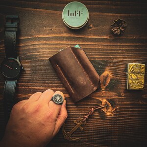 May include: A brown leather wallet with stitching detail, a silver compass ring, a silver lighter with the words "US NAVY" on it, a small round container with the words "lofi: leather goods" on it, and a small wooden anchor charm.