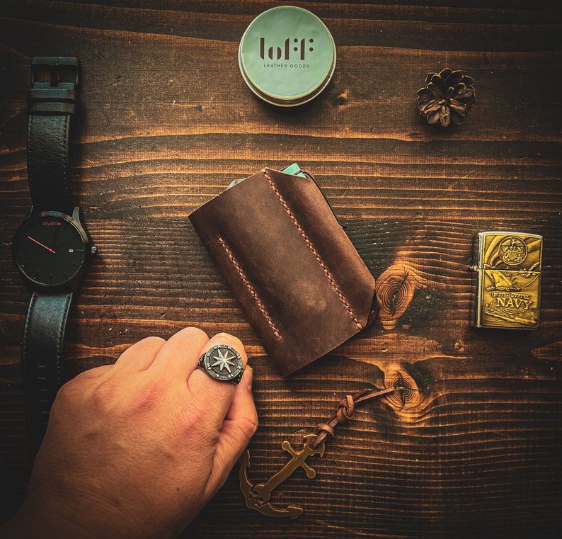 May include: A brown leather wallet with stitching detail, a silver compass ring, a silver lighter with the words "US NAVY" on it, a small round container with the words "lofi: leather goods" on it, and a small wooden anchor charm.