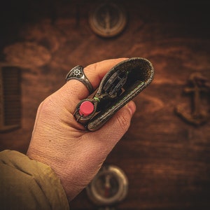 May include: A hand holding a brown leather wallet with a red button and a silver ring on the finger. The wallet is open and the inside is visible.