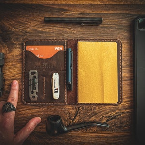 Może przedstawiać: Brązowy sk&oacute;rzany portfel z notesem, kartą kredytową, długopisem i wielofunkcyjnym narzędziem. Portfel ma napis "Large Leather Goods" z przodu.