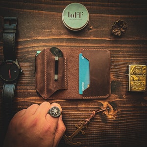 May include: A brown leather wallet with a card slot and a pocket for a knife. The wallet is open and a credit card is visible. The wallet is on a wooden surface with a watch, a small container, a lighter, and a ring.