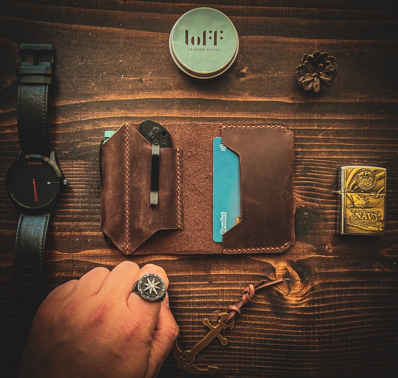 May include: A brown leather wallet with a card slot and a pocket for a knife. The wallet is open and a credit card is visible. The wallet is on a wooden surface with a watch, a small container, a lighter, and a ring.