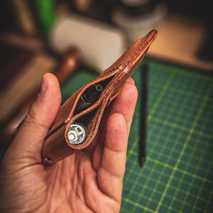 May include: A brown leather pen case with two compartments, one holding a pen and the other a small, silver-coloured object. The case is held in a hand against a blurred background.