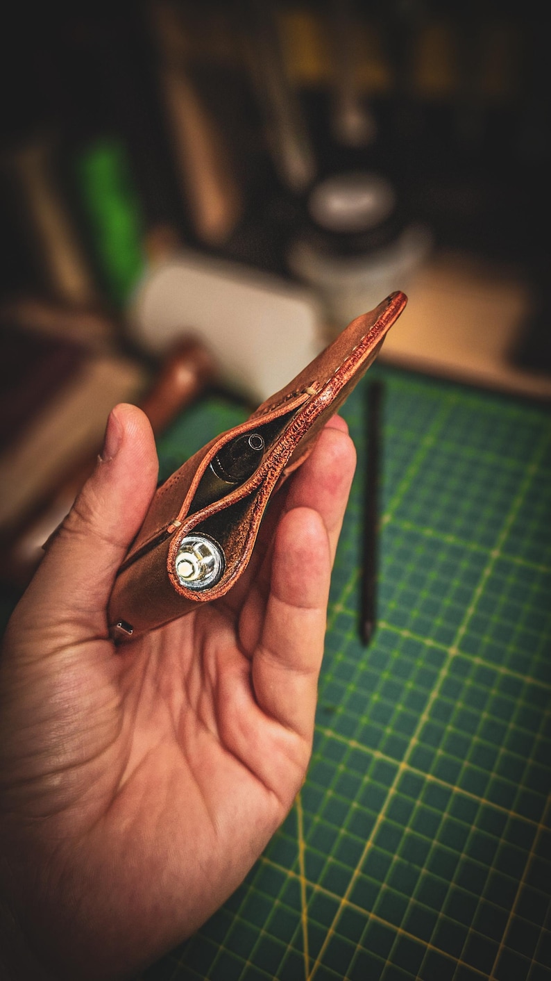 May include: A brown leather pen case with two compartments, one holding a pen and the other a small, silver-coloured object. The case is held in a hand against a blurred background.