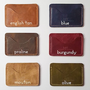 May include: Six leather cardholders in English tan, blue, praline, burgundy, mouton, and olive. Each has a curved pocket and visible stitching. The text "loft LEATHER GOODS" and colour names are printed on each. Measurements are not available.