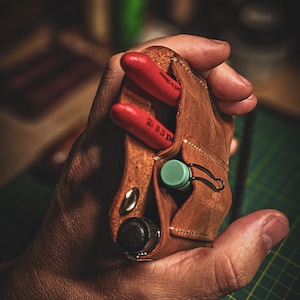 May include: A brown leather tool holder with red tools and a green cap. The holder has a snap closure and a metal loop. The tools are red with white text. The holder is held in a hand.