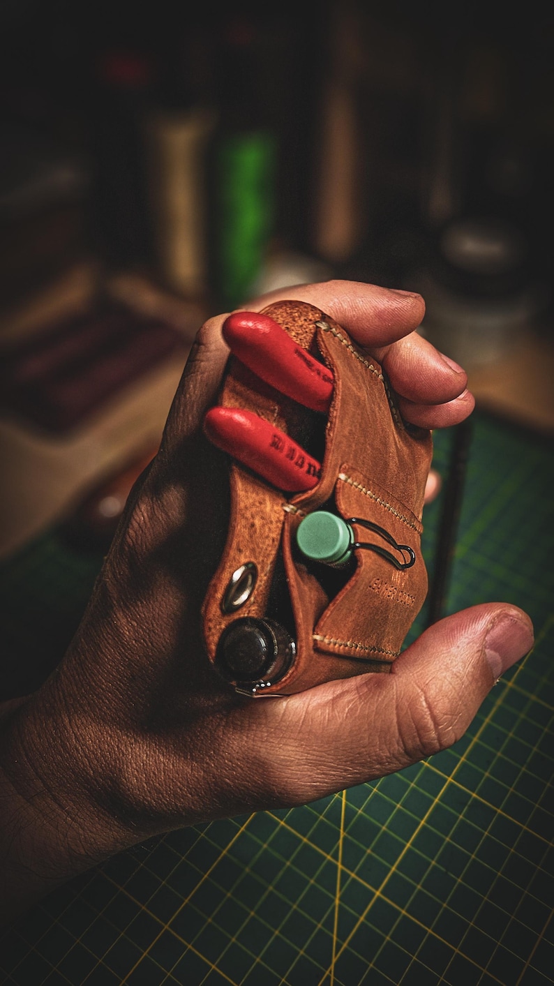 May include: A brown leather tool holder with red tools and a green cap. The holder has a snap closure and a metal loop. The tools are red with white text. The holder is held in a hand.