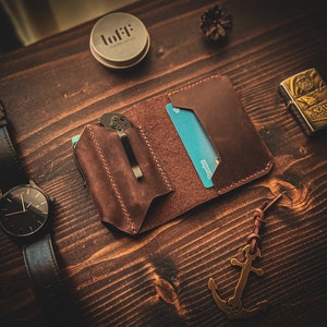 May include: A brown leather wallet with a card slot and a pocket for a knife. The wallet is on a wooden surface with a watch, a small container, and a lighter.