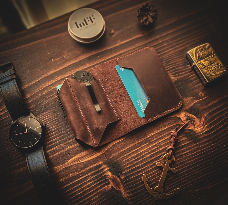 May include: A brown leather wallet with a card slot and a pocket for a knife. The wallet is on a wooden surface with a watch, a small container, and a lighter.