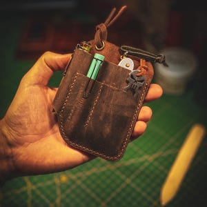 Peut inclure: Un organisateur de poche en cuir marron foncé doté de deux fentes verticales, d'une boucle en cuir sur le dessus et de coutures apparentes. L'organisateur contient un stylo vert clair et une breloque en métal en forme de crâne.