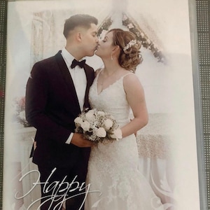 May include: A wedding photo of a couple kissing. The bride is wearing a white lace dress and the groom is wearing a black suit. The photo is set against a white background with the text "Happy Anniversary 10.12.2019" written below the couple.