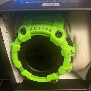 May include: A bright neon green digital watch with a round black face and a thick, rugged bezel. The watch features the brand name ACTIVA printed on the top, with buttons labeled LIGHT, START, and RESET on the sides. It is housed in a dark box.