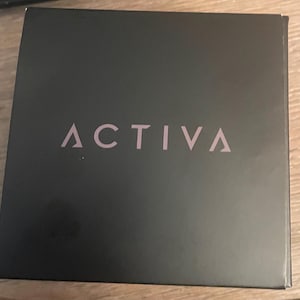 May include: A square black box with the word ACTIVA printed in a light pink, sans-serif font across the center. The box has a smooth, matte finish and features a clean, minimalist design with the brand name clearly visible on the front surface.
