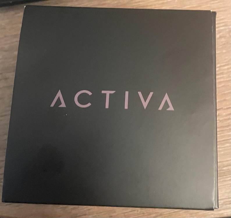 May include: A square black box with the word ACTIVA printed in a light pink, sans-serif font across the center. The box has a smooth, matte finish and features a clean, minimalist design with the brand name clearly visible on the front surface.