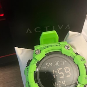 May include: A bright neon green digital watch with a round black face displaying the time 1:59, the date 4-24, and the day MON. The bezel features the text ACTIVA, LIGHT, MODE, START, RESET, and WATER RESIST with four visible silver-toned screws.