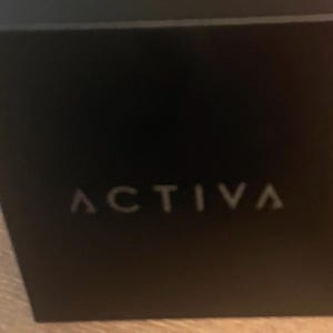 May include: A square black box with the word ACTIVA printed in a light gray, sans-serif font centered on the front face. The box has a smooth, matte finish and sharp, clean edges, presenting a simple and uniform appearance throughout the object.