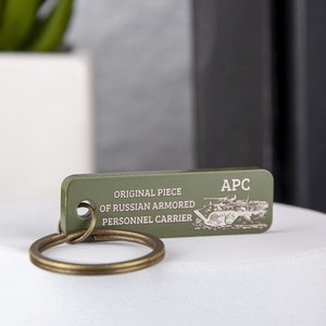 Pode incluir: Um chaveiro de metal verde com uma pequena imagem detalhada de um veículo blindado de transporte de pessoal russo. O chaveiro tem o texto "ORIGINAL PIECE OF RUSSIAN ARMORED PERSONNEL CARRIER" e "APC".