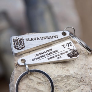 May include: A silver keychain with two metal tags. The top tag has the Ukrainian trident symbol and the text "SLAVA UKRAINI". The bottom tag has the text "T-72 ORIGINAL PIECE OF RUSSIAN TANK SKIN RECYCLED IN UKRAINE 2022".