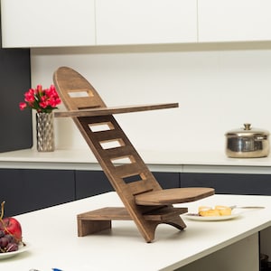 Puede incluir: Un soporte de madera con una superficie inclinada y un pequeño estante. El soporte está hecho de madera marrón oscura y tiene un acabado natural. Está diseñado para sostener una computadora portátil o una tableta.