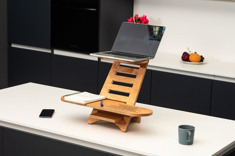 May include: A wooden laptop stand with a notebook and pen on the top shelf. The stand is on a white countertop with a grey mug and a mobile phone.