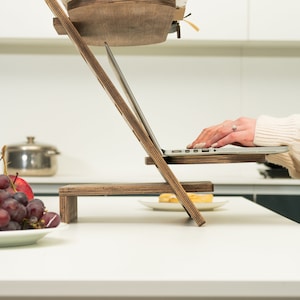 May include: A wooden laptop stand with two levels, a laptop on the top level, and a person's hands typing on the keyboard. The stand is on a white countertop with a plate of fruit and a plate of food on the lower level.