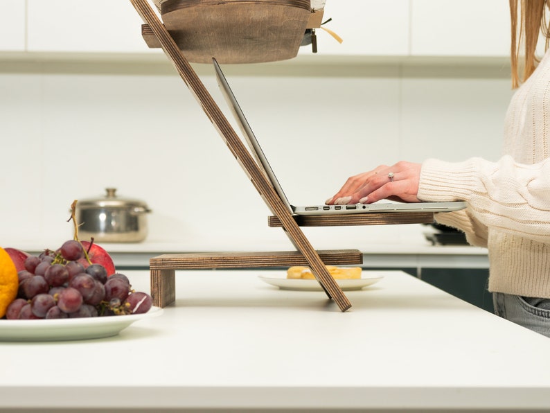 May include: A wooden laptop stand with two levels, a laptop on the top level, and a person's hands typing on the keyboard. The stand is on a white countertop with a plate of fruit and a plate of food on the lower level.