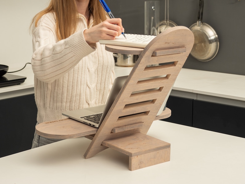 May include: A wooden laptop stand with a built-in shelf for a notebook. The stand is light brown and has a natural wood grain finish. The stand is angled to provide ergonomic support for a laptop.