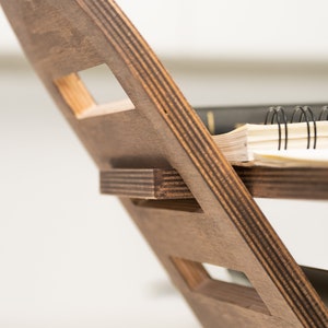 May include: A wooden magazine rack with a dark brown stain. The rack has a unique design with multiple slots for holding magazines or books. A notebook with a black spiral binding and a blue pen are resting on the top shelf.