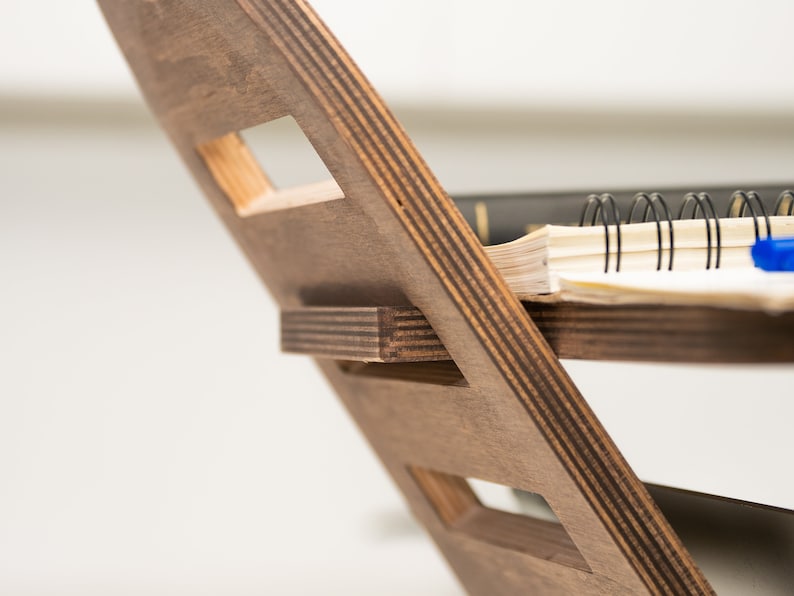May include: A wooden magazine rack with a dark brown stain. The rack has a unique design with multiple slots for holding magazines or books. A notebook with a black spiral binding and a blue pen are resting on the top shelf.