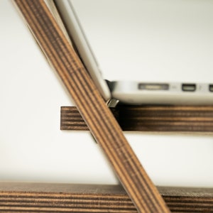 May include: A wooden laptop stand with a dark brown stain. The stand is made of plywood and has a simple design. The stand is designed to elevate a laptop to a comfortable viewing angle.