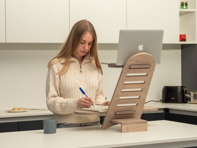 May include: A person working at a standing desk with a laptop and a notebook. The desk is made of wood and has a built-in shelf for the laptop. The person is wearing a white sweater and is writing in the notebook with a blue pen.