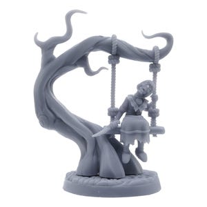 May include: A gray miniature sculpture of a girl on a swing attached to a tree. The girl is wearing a dress and holding a knife. The tree has a twisted trunk and branches. The sculpture is on a round base.