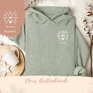 Puede incluir: Una sudadera con capucha de color verde claro con un diseño gráfico blanco en la parte delantera. El diseño presenta un corazón y las letras "ANT" y "TE" dispuestas en forma de cruz. El texto "EST. 2024" está debajo del diseño.